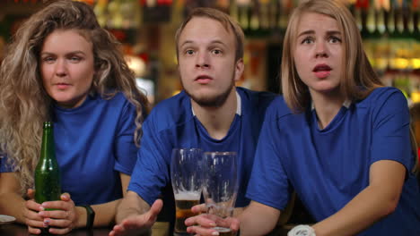 Close-up-group-of-fans-emotionally-watching-a-sports-broadcast-in-a-bar-on-TV-with-beer-men-and-women-in-blue-T-shirts-of-different-races-are-upset-after-losing-and-losing-their-team.